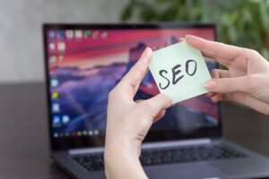 In front of a laptop, hands hold a sticky note that says SEO.