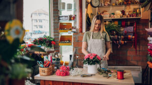 Graceful female florist making a beautiful bouquet in a round box while wearing an apron and working in a flower shop. Florist workplace. Small business concept.